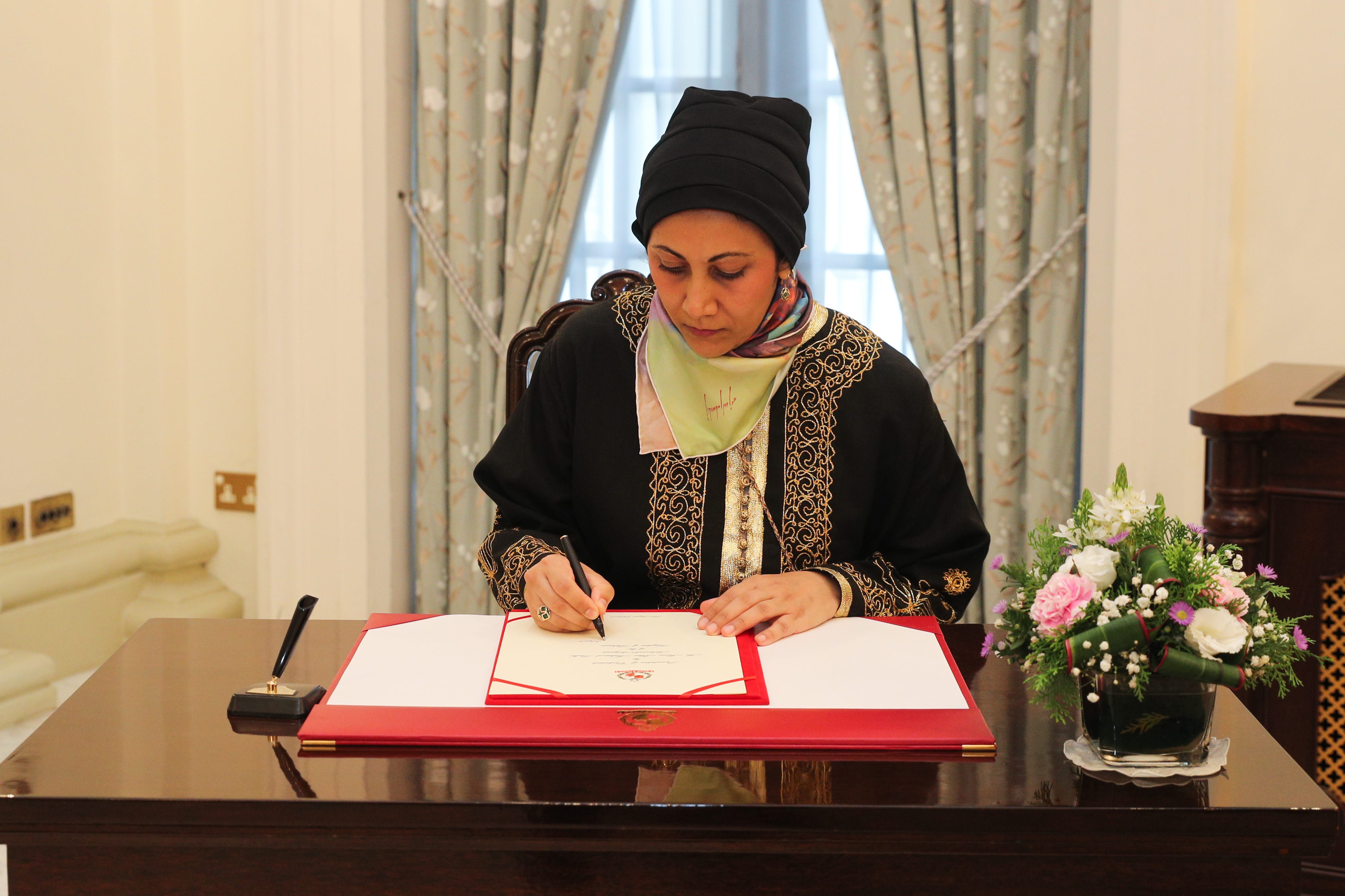 Woman in black head covering, signing document with seal at wooden desk.